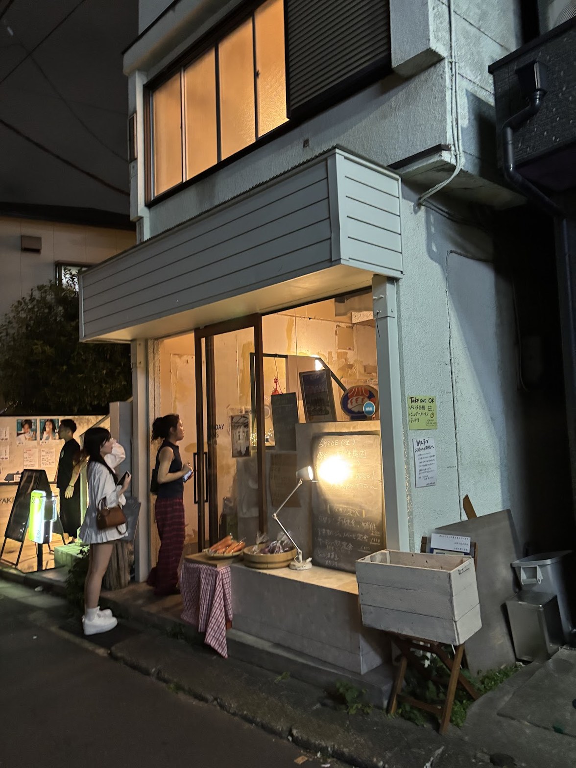 夜の路地に面した店舗外観、ガラス窓から温かい光が漏れ、手書き黒板と野菜が店先に並ぶ