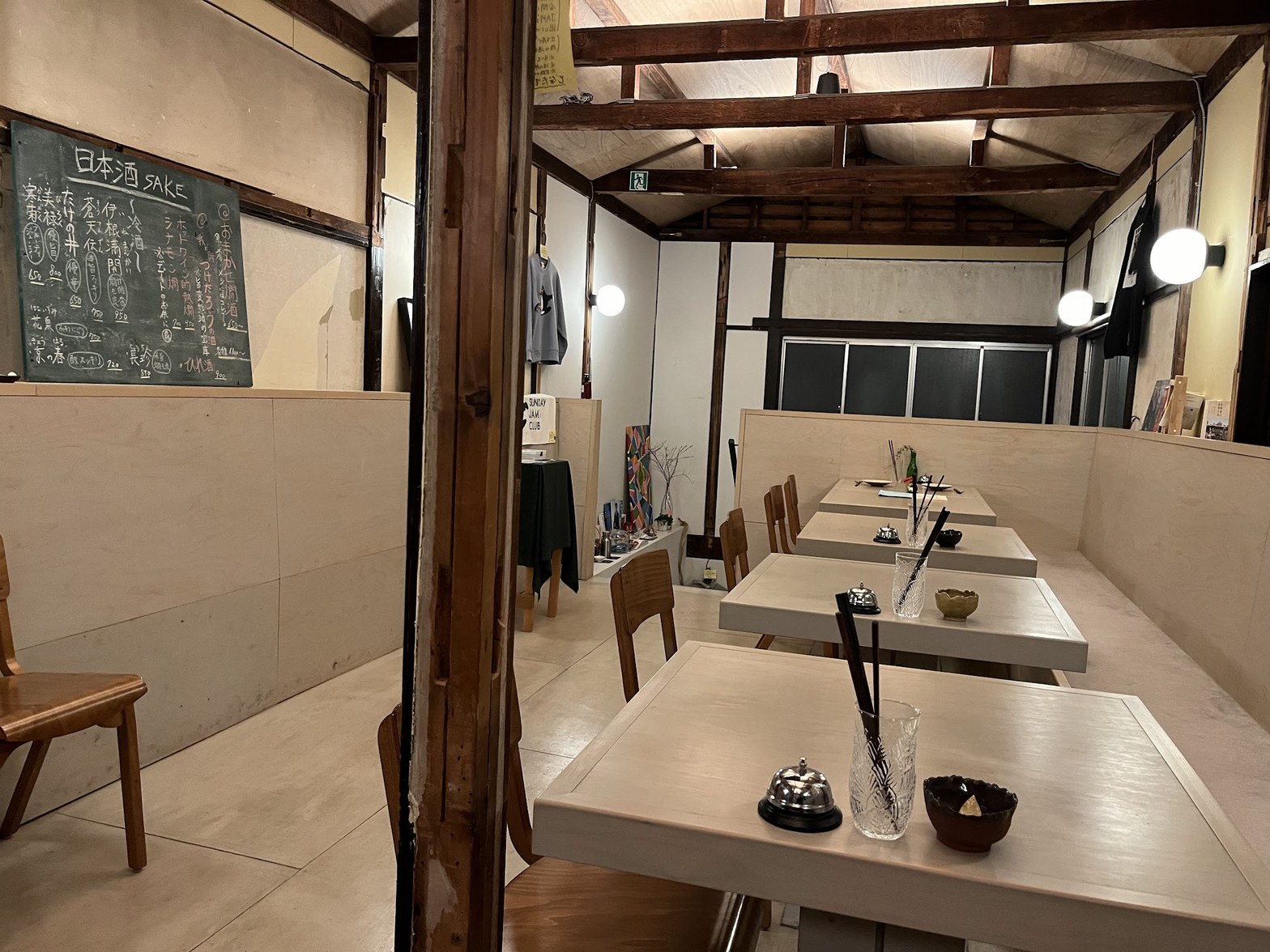 黒板に「日本酒 SAKE」と銘柄が手書きされた夜の店内、白木テーブルにベルと箸立てが並ぶ