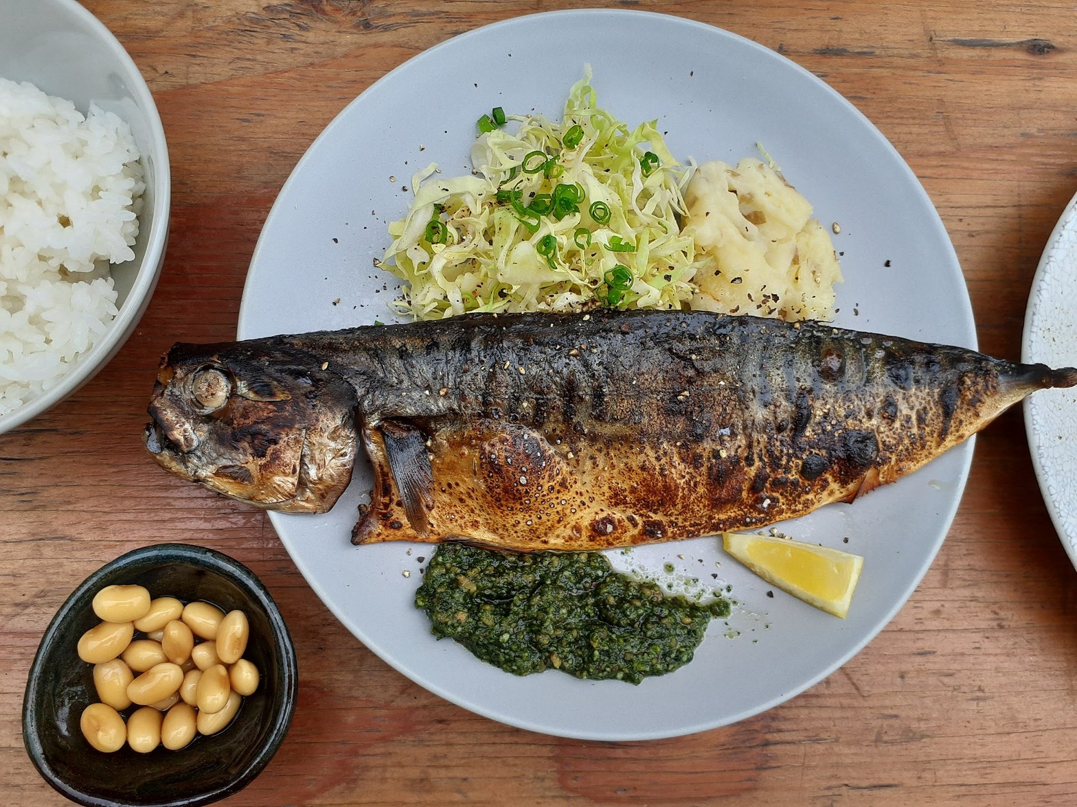 グレーの丸皿に丸ごと一尾の焼き魚、大葉ジェノベーゼとレモン添え、左下に黒い小鉢と白米